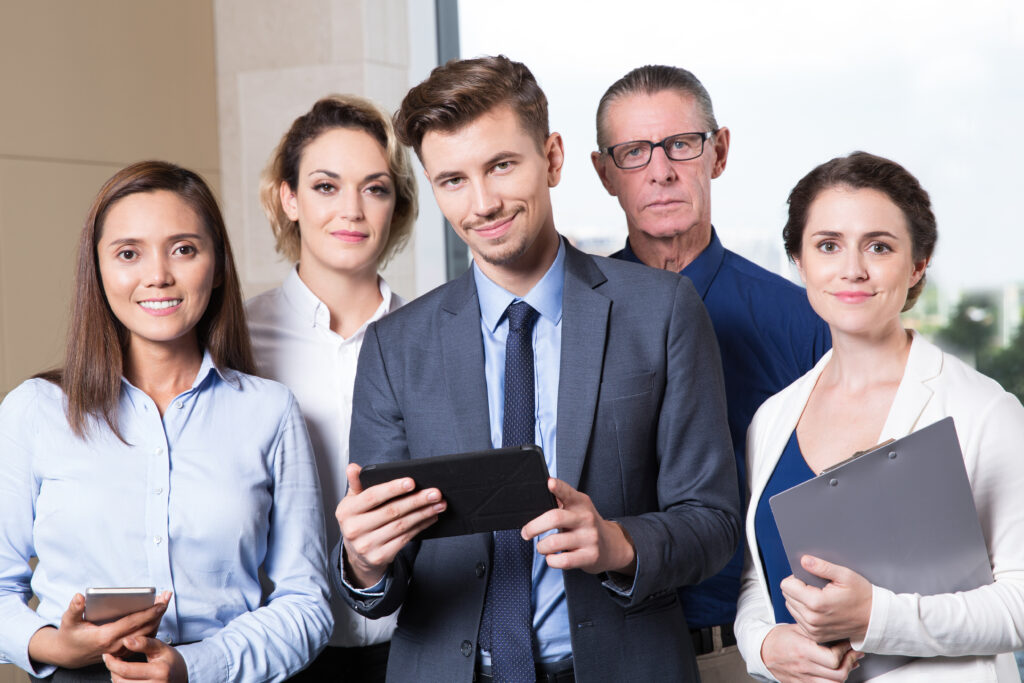 Group of five successful business people standing in conference room, looking at camera. Young businessman holding tablet computer, businesswoman with clipboard, her Asian colleague with smartphone